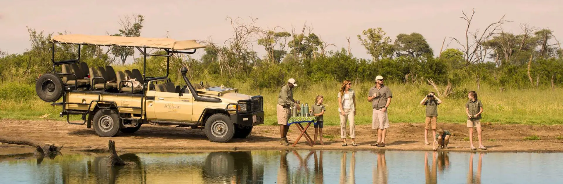 A family enjoys sundowner drinks with their guide next to a body of water in the foreground | Go2Africa
